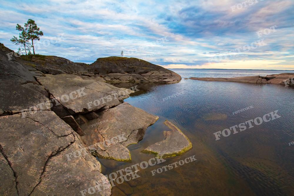 На фотографии запечатлен каменистый берег в Карелии. Неровные, гладко выветренные скалы спускаются к воде, создавая естественные лестницы и платформы. Несколько одиноких деревьев растут прямо из камня, их корни ухитряются найти питание в щелях скал. Над ландшафтом раскинулось облачное небо, которое отражается в тихой воде, придавая сцене мирный характер. Этот вид может использоваться для иллюстрации тем, связанных с природой, в туристических брошюрах и в качестве вдохновения для художественных работ.